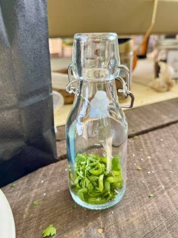 A small glass bottle with a metal clasp contains chopped green herbs, sitting on a wooden surface.