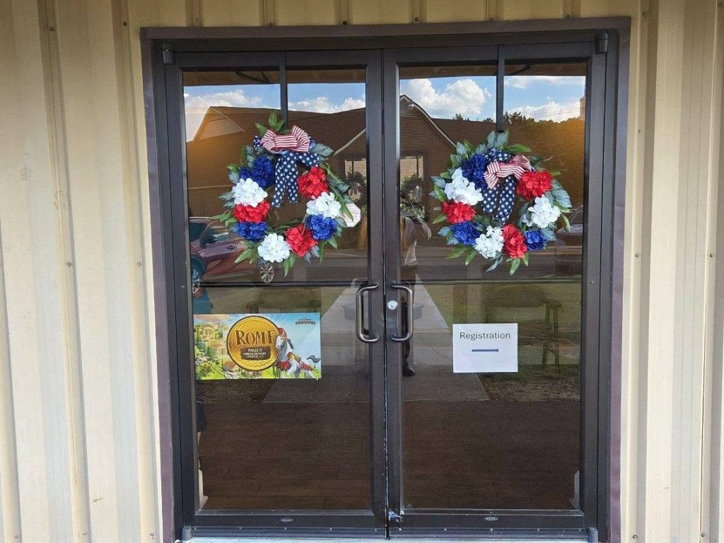 Glass double doors with patriotic wreaths, a "Registration" sign, and a poster for a Rome-themed event, reflecting a building and cars outside.