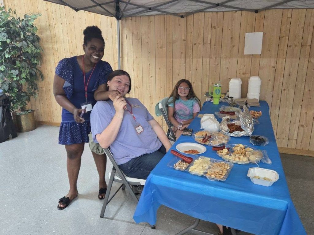 Three people smiling near a table covered with a blue cloth, featuring various snacks and food trays, indoors with wooden walls in the background.