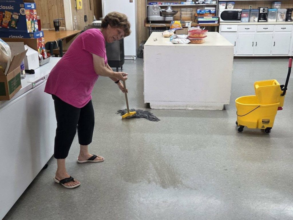 A woman wearing a pink shirt and black pants is mopping the floor in a kitchen with a yellow mop bucket nearby.