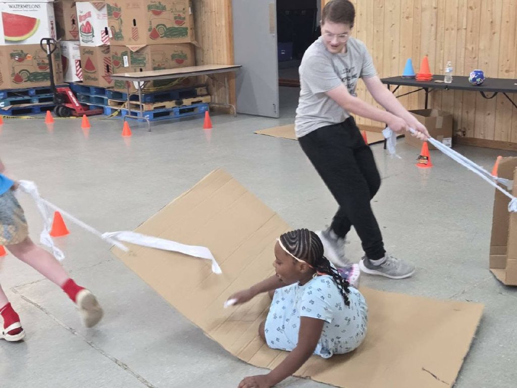Children playing indoors, pulling a girl seated on a large piece of cardboard with strips of cloth, surrounded by orange cones and cardboard boxes.
