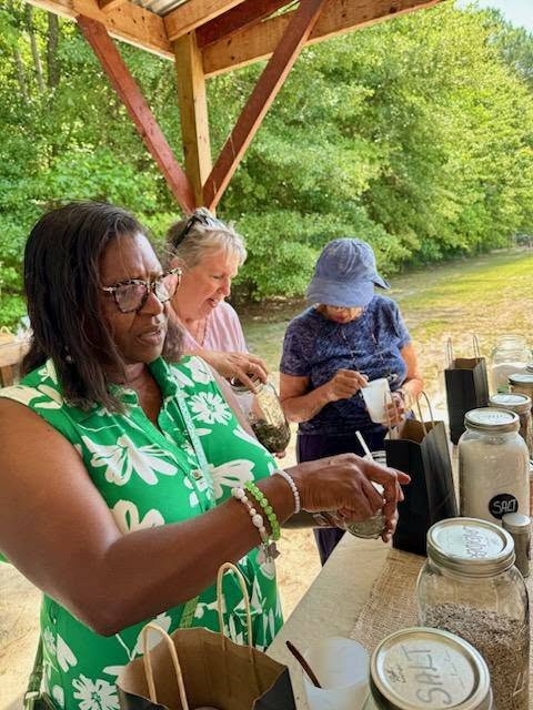 Three women outdoors fill cups from large labeled jars on a wooden table, under a shelter, with greenery in the background.