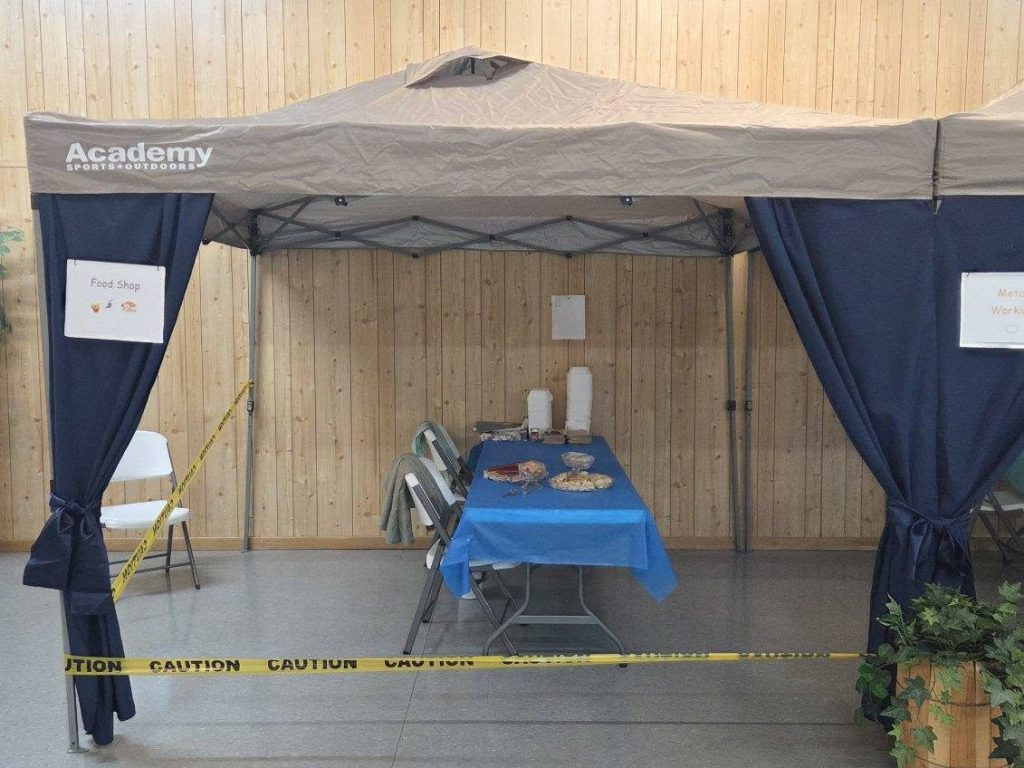 A food booth under a canopy with a table covered in snacks, chairs, caution tape, and signs reading "Food Shop" and "Academy Sports + Outdoors."