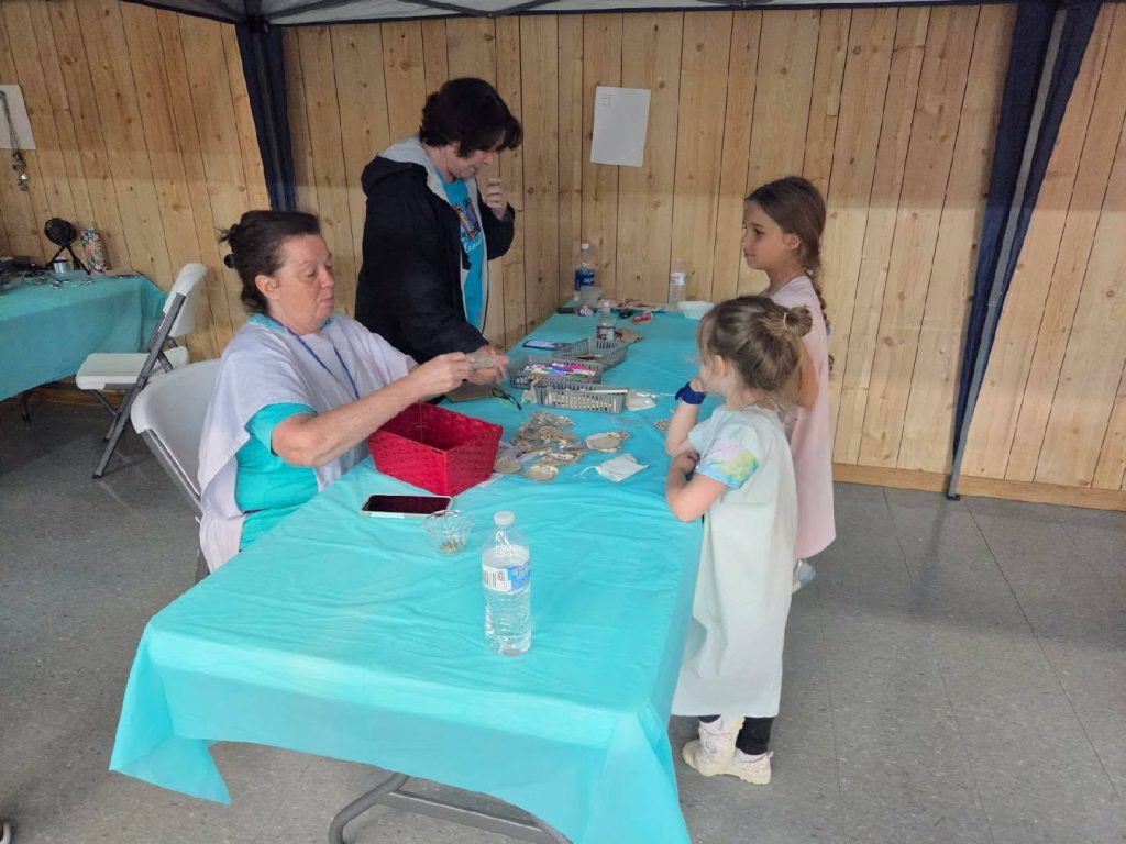 Two women behind a table with crafts, talking to two young girls in a room with wooden walls and blue tablecloths.