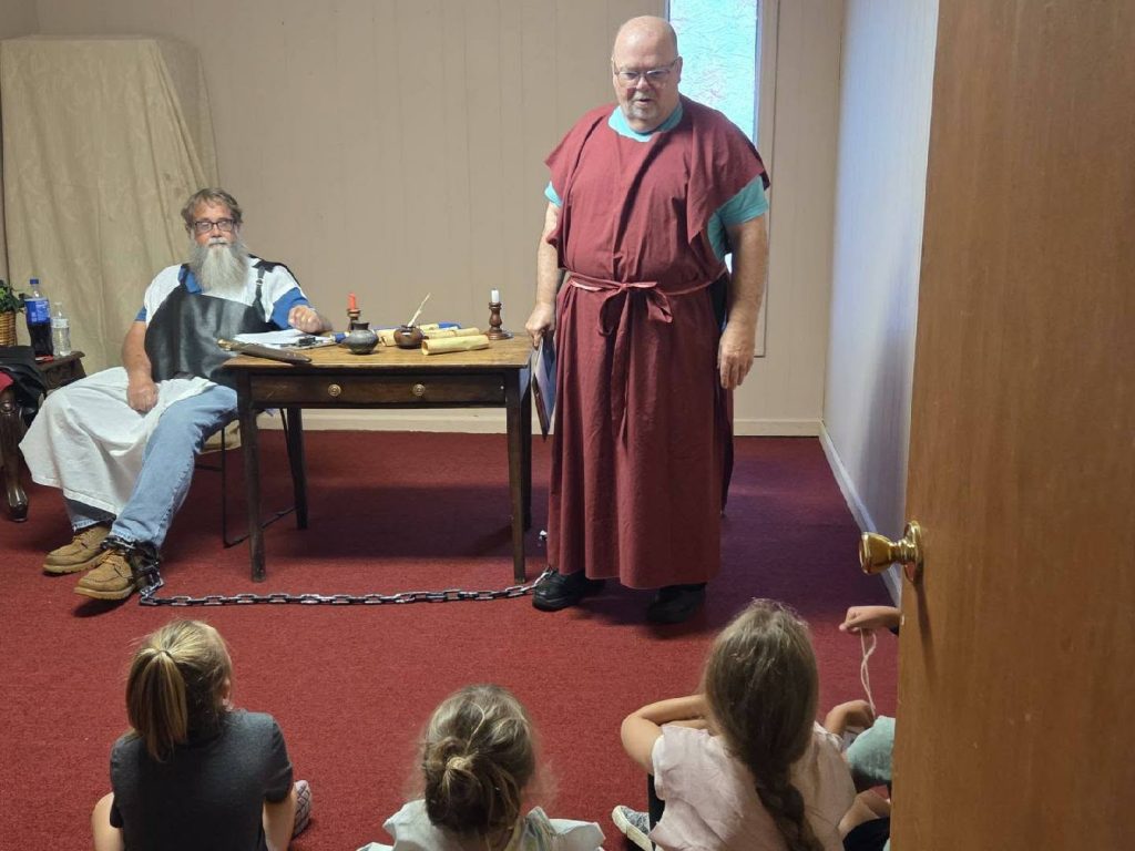 Two men in costume reenact a historical scene for children seated on the floor, one man is chained and sitting at a desk with scrolls.