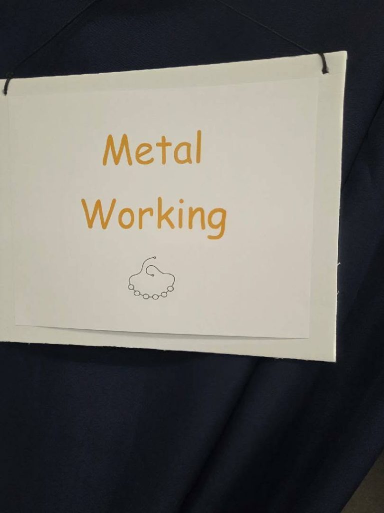 A sign reading "Metal Working" in orange letters, hanging on a dark background, with a simple line drawing of a wire and beads below the text.