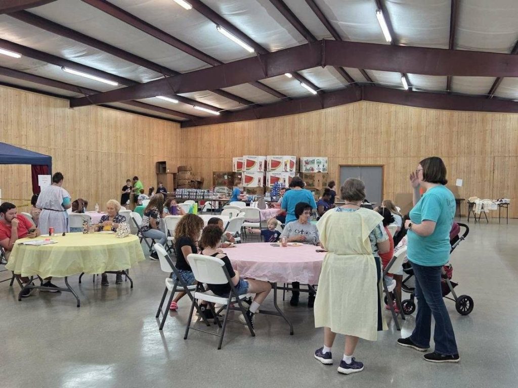 People of various ages gather at round tables in a large hall, engaging in conversation and activities. Some stand while others sit, creating a lively community atmosphere.