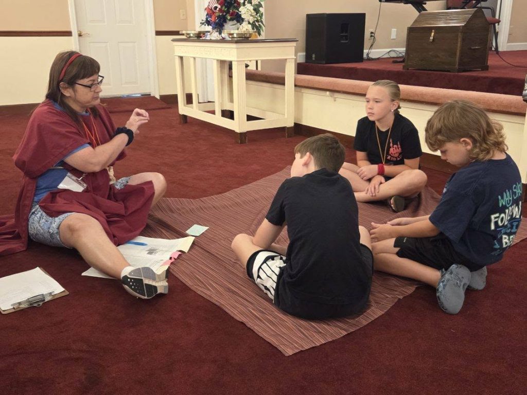 A woman sits on the floor leading a small group of three children in discussion inside a room with red carpet and a low stage.