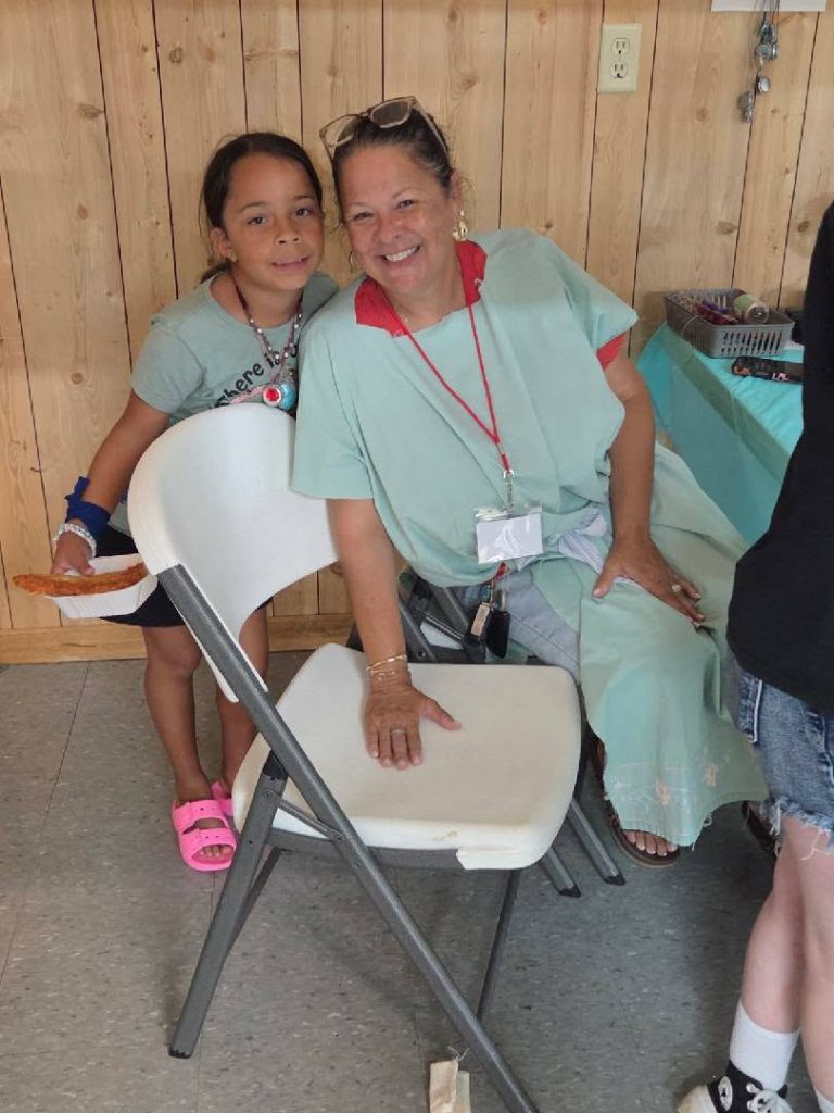A smiling woman sitting on a chair and a young girl standing beside her, inside a room with wooden walls.