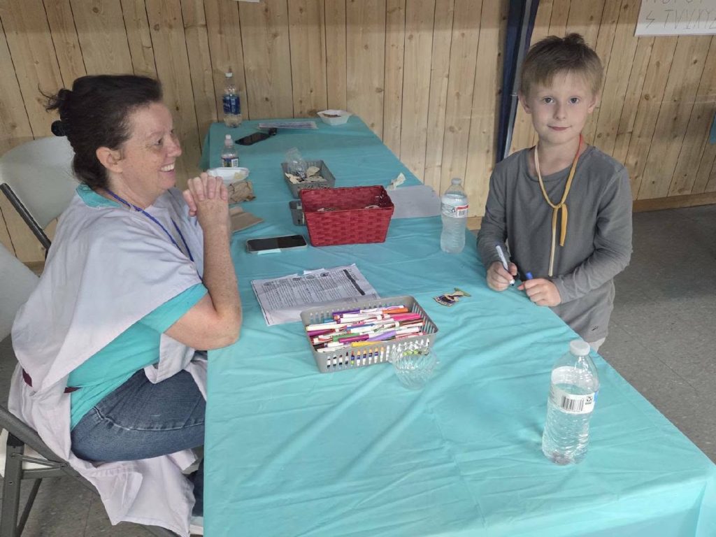 A smiling woman sits at a table with a young child, art supplies, water bottles, and papers on a blue tablecloth in a wood-paneled room.