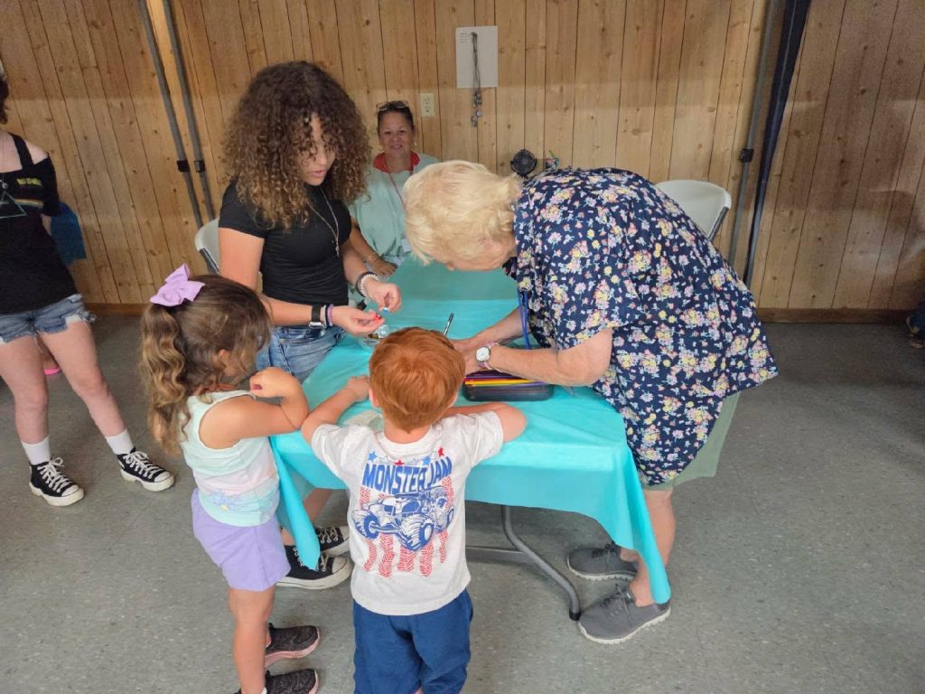 elderly woman signs something at a table while children and a young woman watch, inside a room with wood-paneled walls.