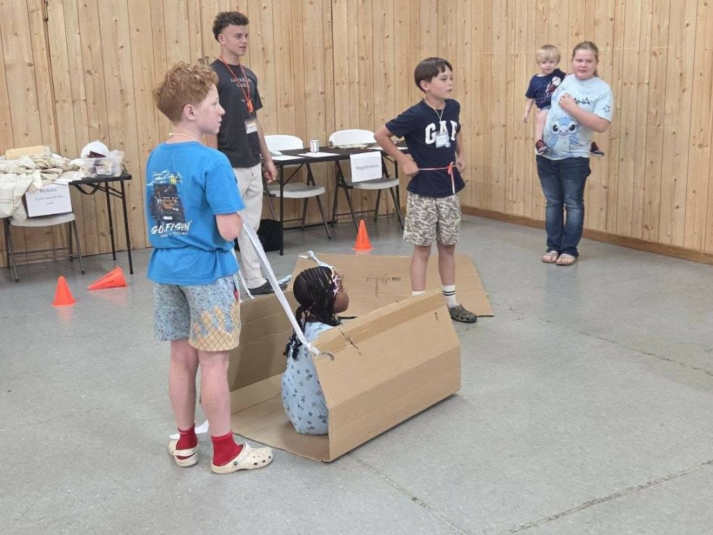 Children play with cardboard boxes indoors while adults and another child observe nearby, in a room with a wooden wall and orange cones on the floor.