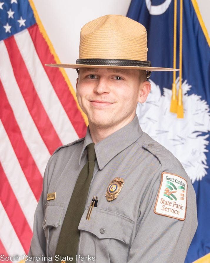 A South Carolina State Park ranger in uniform stands in front of the American and South Carolina flags, smiling for an official portrait.
