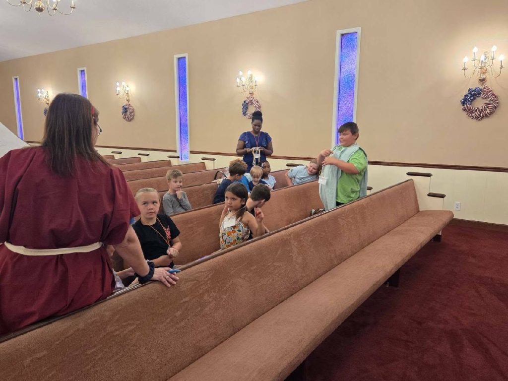 A group of children and two adults sit and stand on church pews, engaging in conversation inside a softly lit room with stained glass windows.