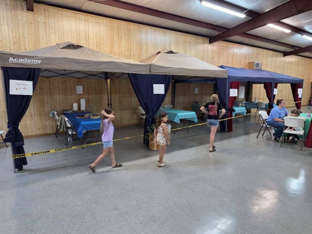 Children walk past indoor booths in a community center, with tables, signs, and caution tape sectioning off areas. People are seated at tables near the booths.