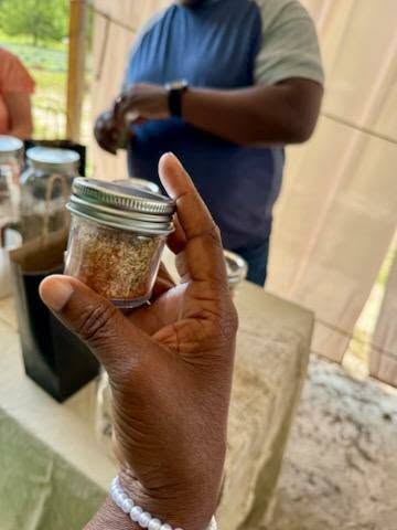 A hand holding a small jar with spices; another person stands in the background at a table with jars and containers.