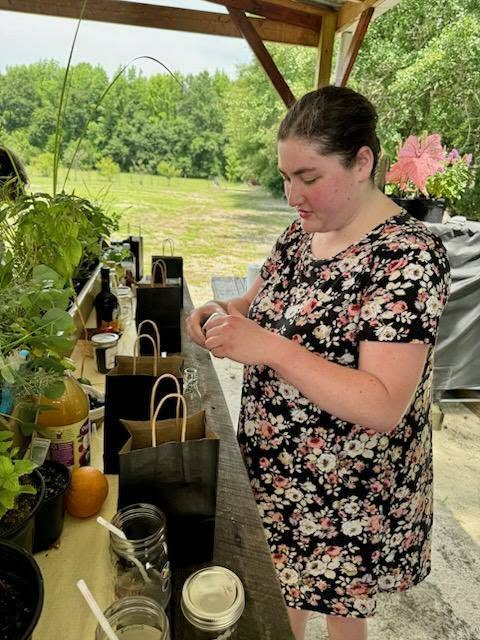 A woman in a floral dress prepares items on an outdoor table with jars, bags, plants, and bottles under a wooden shelter surrounded by greenery.