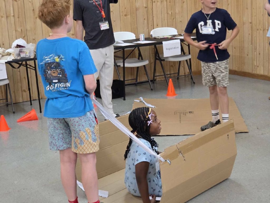 Three children play indoors with cardboard and streamers; one girl sits in a cardboard box while two boys stand nearby. Orange cones and tables are in the background.