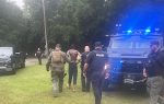 Police officers escort a handcuffed, shirtless man past police vehicles and an armored truck near a wooded area.