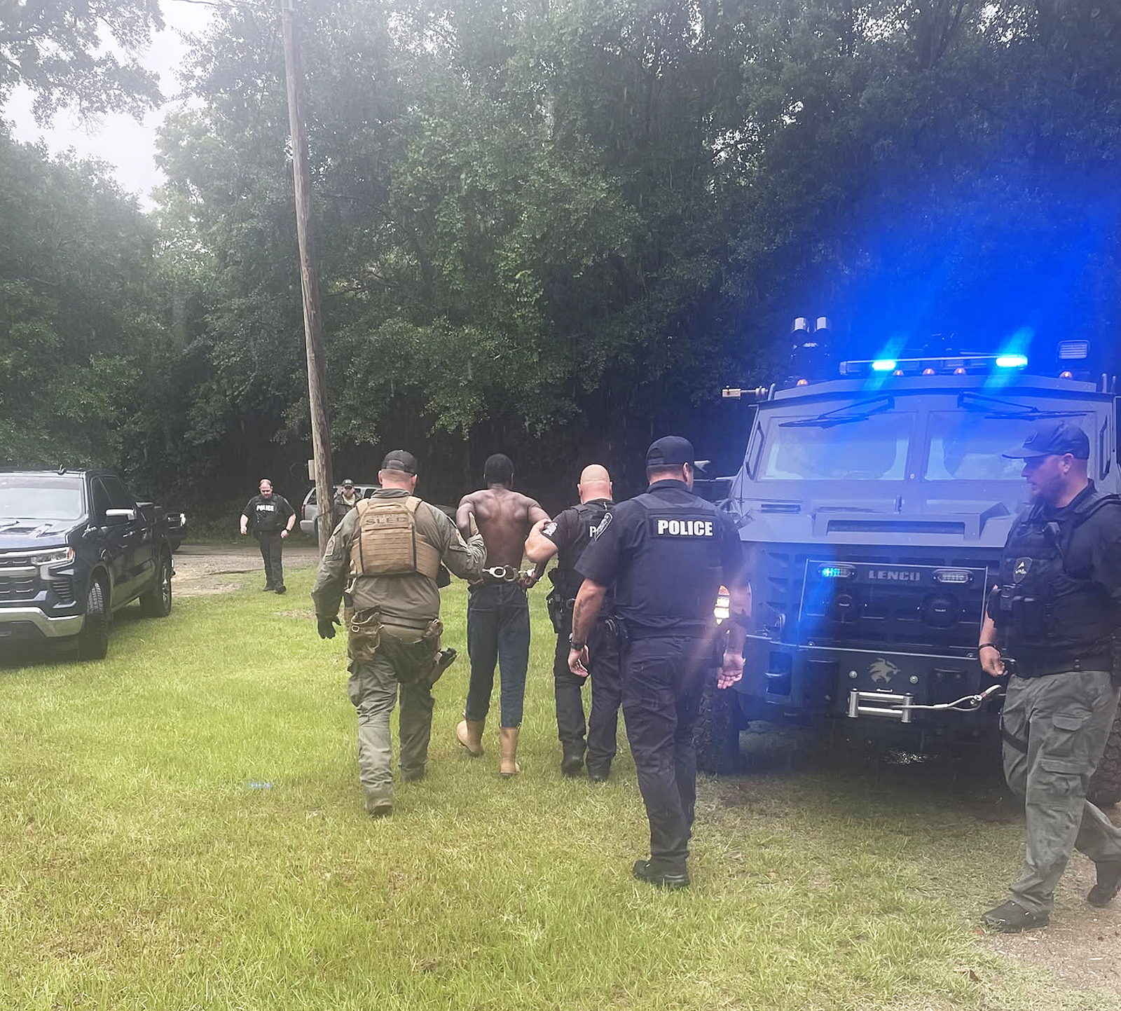 Police officers escort a handcuffed, shirtless man past police vehicles and an armored truck near a wooded area.