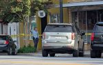 A police officer stands behind yellow caution tape near parked cars and storefronts on a city street, partially shaded by a tree.