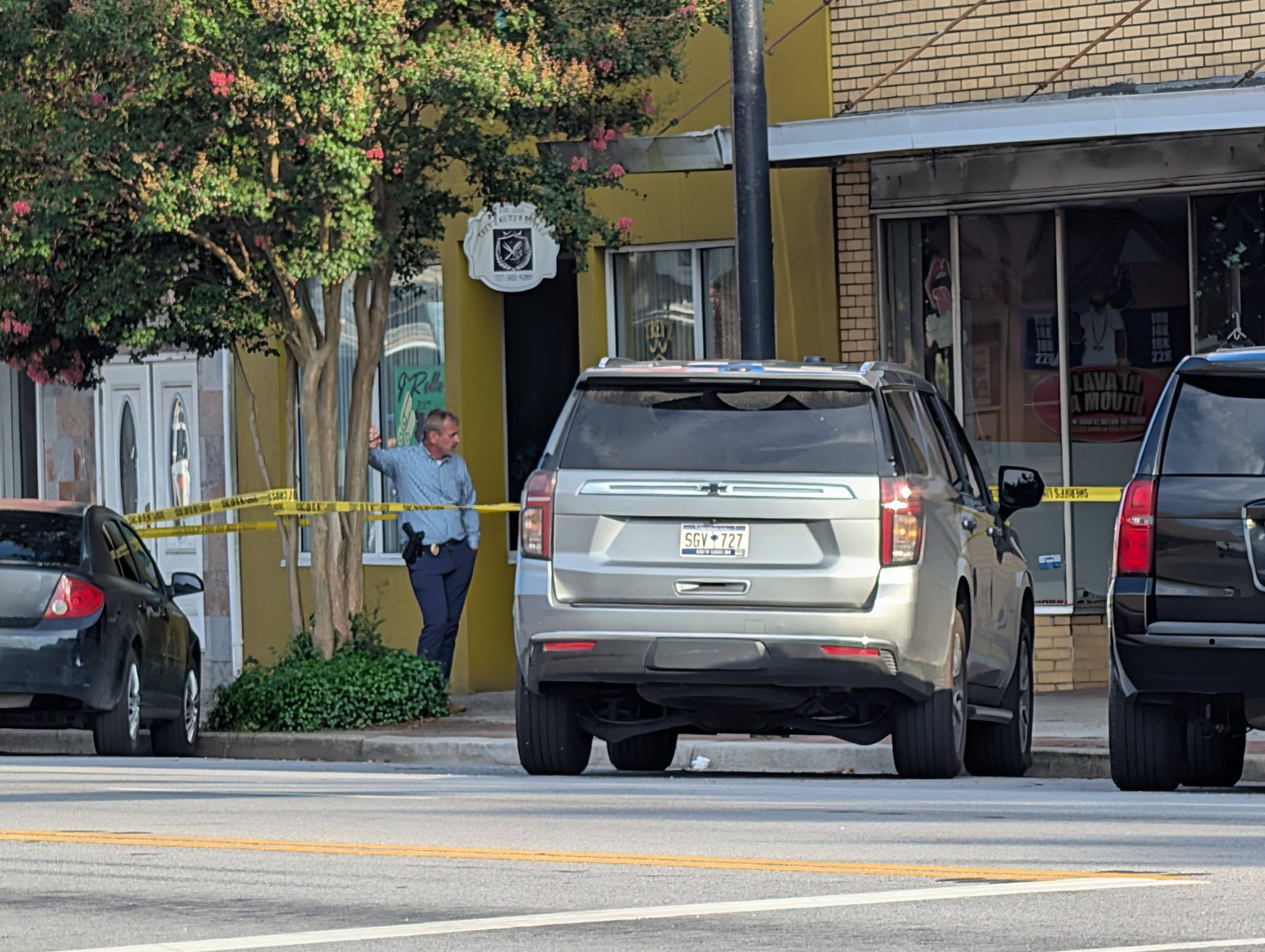 A police officer stands behind yellow caution tape near parked cars and storefronts on a city street, partially shaded by a tree.