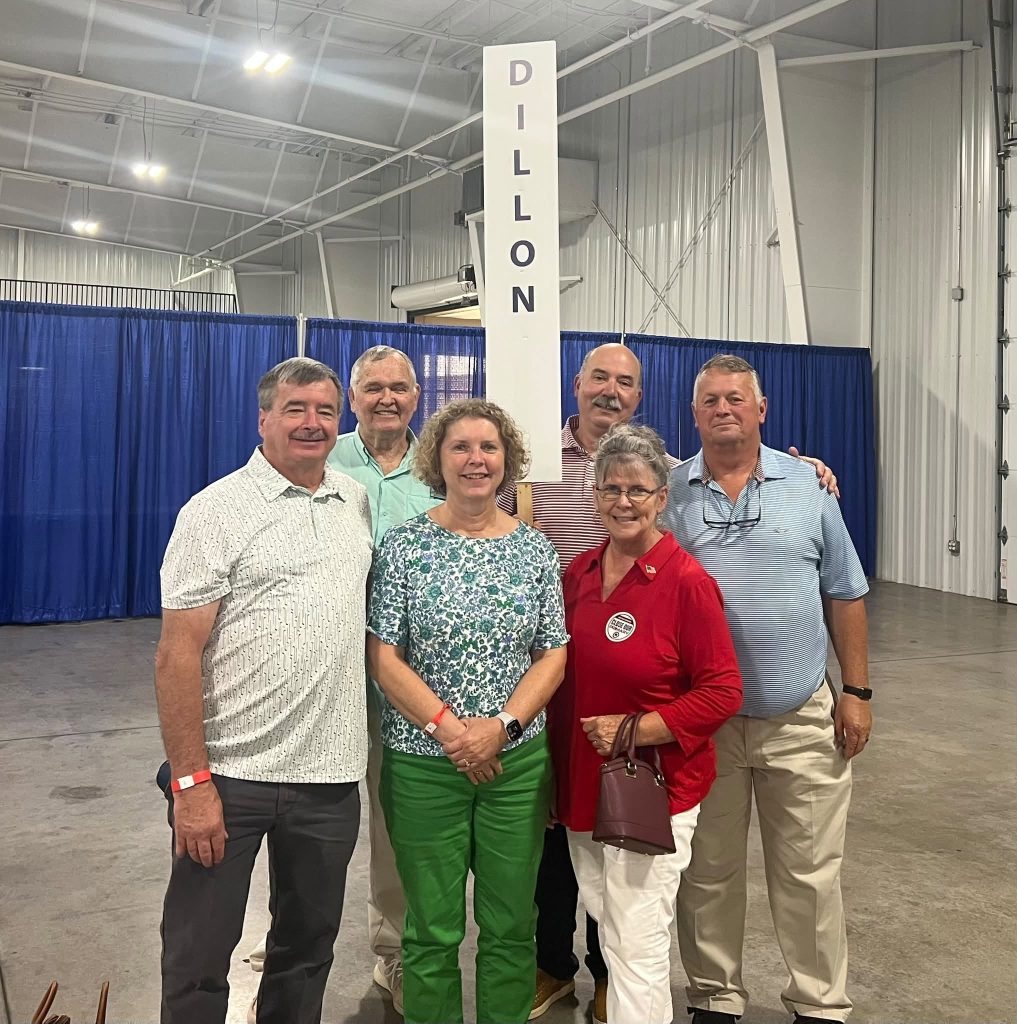 Six adults stand together indoors beside a sign reading "Dillon," smiling for a group photo at what appears to be an event or convention.