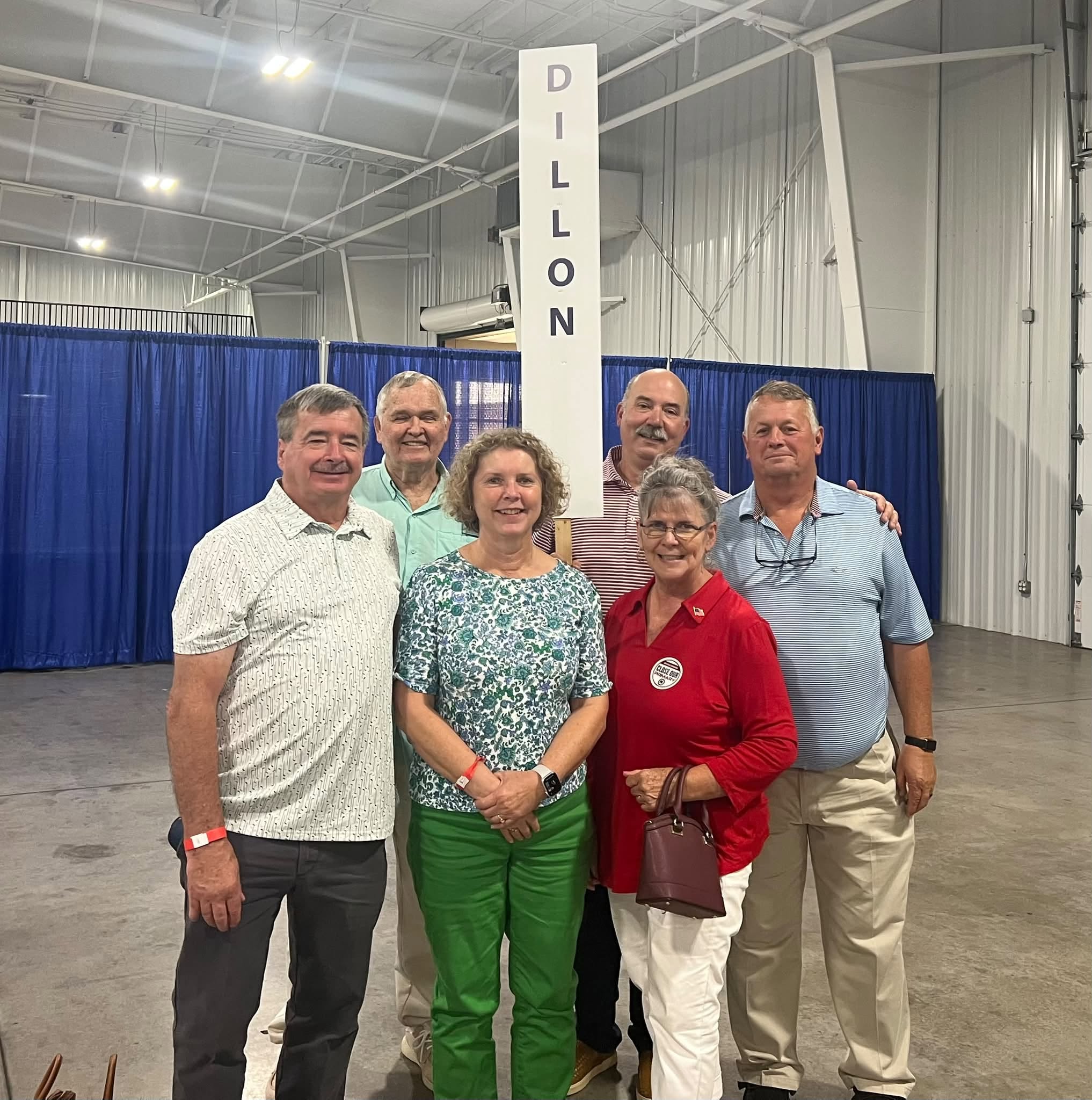 Six adults stand together indoors beside a sign reading "Dillon," smiling for a group photo at what appears to be an event or convention.