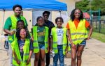 A group of smiling people, adults and children, wearing yellow safety vests, stand outdoors in front of a blue tent on a sunny day.