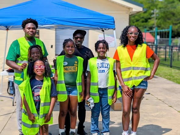 A group of smiling people, adults and children, wearing yellow safety vests, stand outdoors in front of a blue tent on a sunny day.