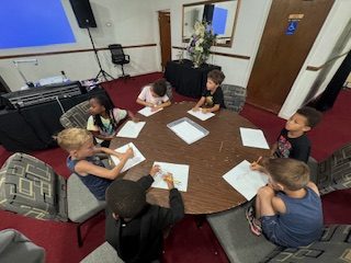 Six children sit around a round table drawing on sheets of paper in a carpeted room with red floors and some equipment visible in the background.