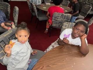 Two children sitting at a table, smiling and holding colorful items, with other children and chairs visible in the background.