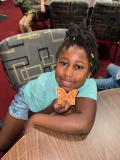 Smiling child sitting at a table, holding a butterfly-shaped cracker, wearing a light blue shirt and jean shorts, with chairs and red carpet in the background.