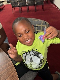Smiling young boy in a neon green shirt holds up a small purple item, sitting at a table with red carpet and chairs in the background.