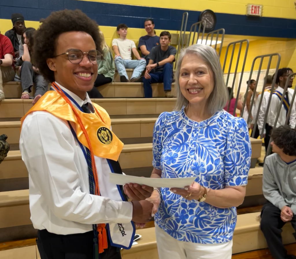 A graduate in a cap and gown smiles while receiving a certificate from a woman in a blue patterned shirt at a school ceremony.