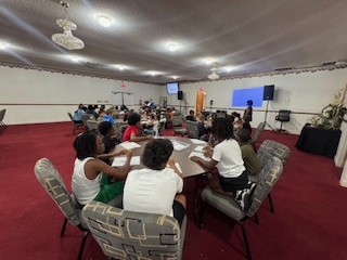 Children sit at round tables in a large room with red carpet, listening to an adult speaker at the front near a projection screen.