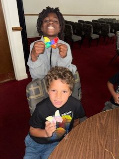 Two children smiling and holding colorful paper crafts while sitting in a room with red carpet and rows of chairs.