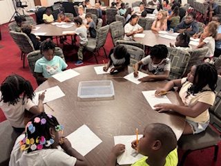 Children sitting at round tables in a classroom, writing on sheets of paper, with focused expressions.
