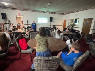 A group of children sit around tables in a room, listening to a presenter at the front near a screen displaying a presentation.