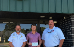 Three men stand in front of a building entrance, one holding a check, under a sign with the letters "BB."