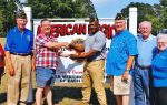 A group of people, some in American Legion caps, stand in front of an American Legion sign while two men in the center shake hands.