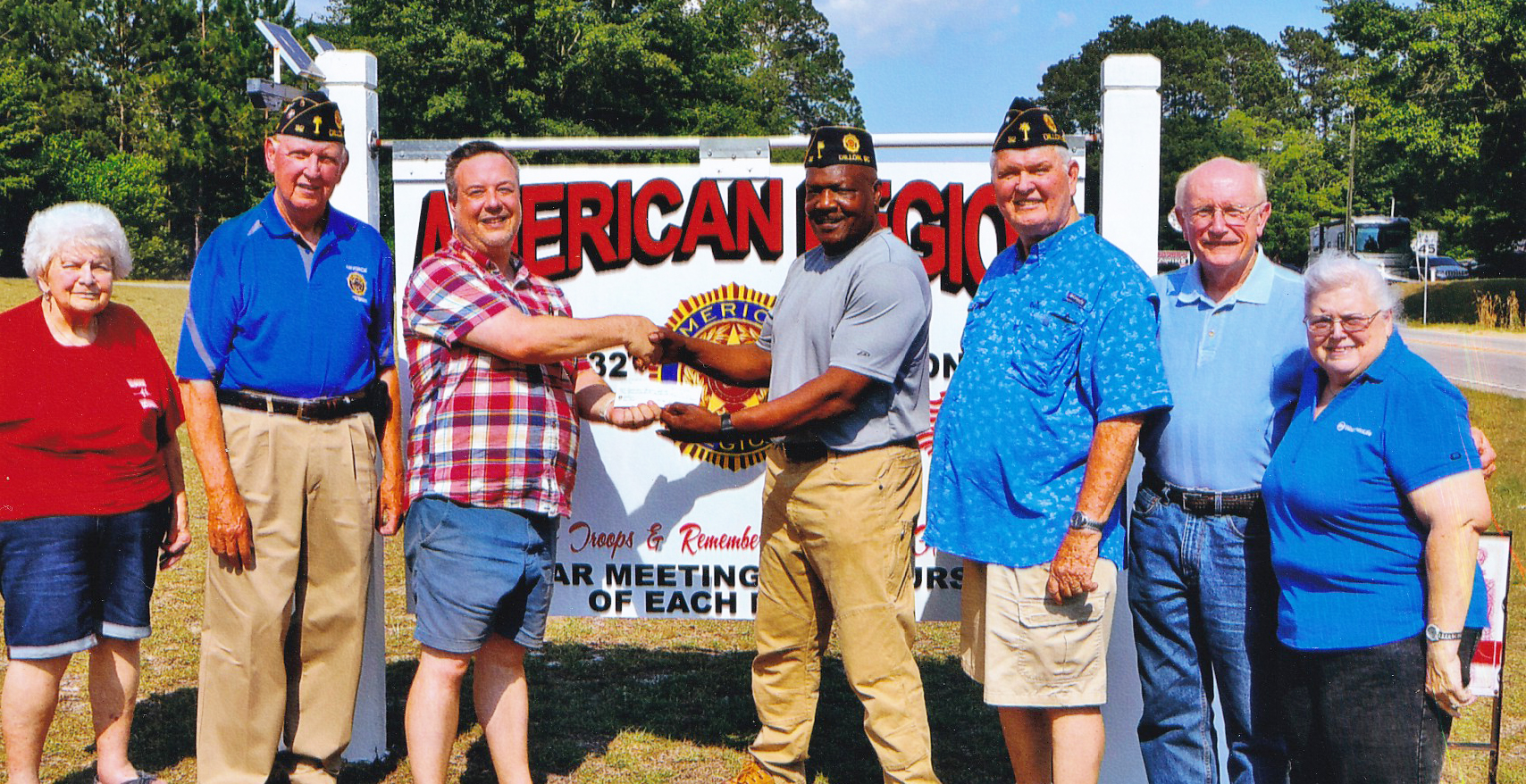 A group of people, some in American Legion caps, stand in front of an American Legion sign while two men in the center shake hands.