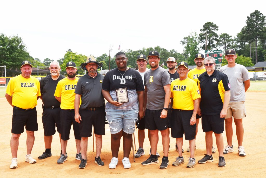 A group of men and one woman standing on a baseball field, with the woman holding a plaque, all smiling at the camera.