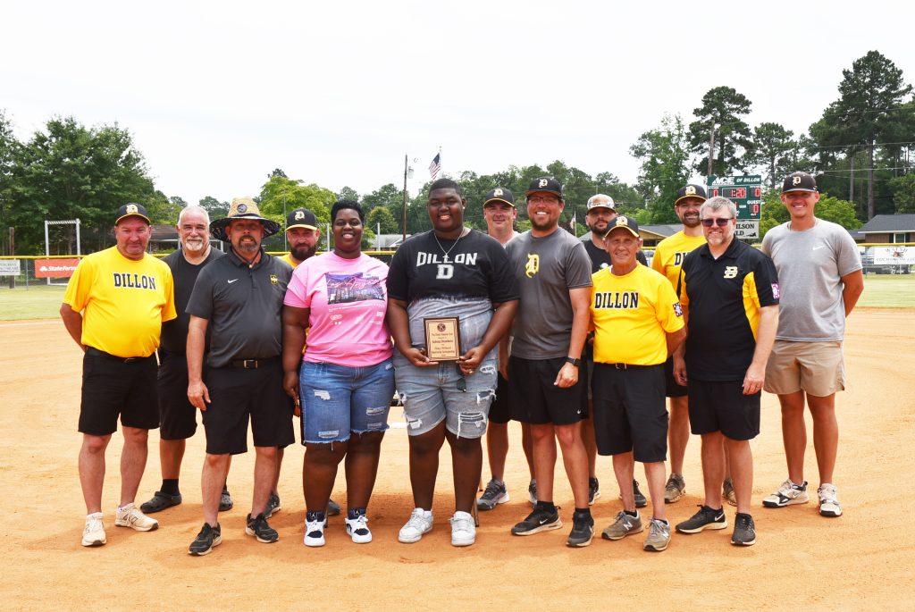 A group of people stand on a baseball field, with one person holding a plaque, posing for a group photo, some wearing sports uniforms.