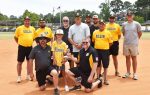 A youth baseball team and coaches pose on a field, some kneeling and some standing, all wearing matching yellow and black uniforms, smiling for the photo.