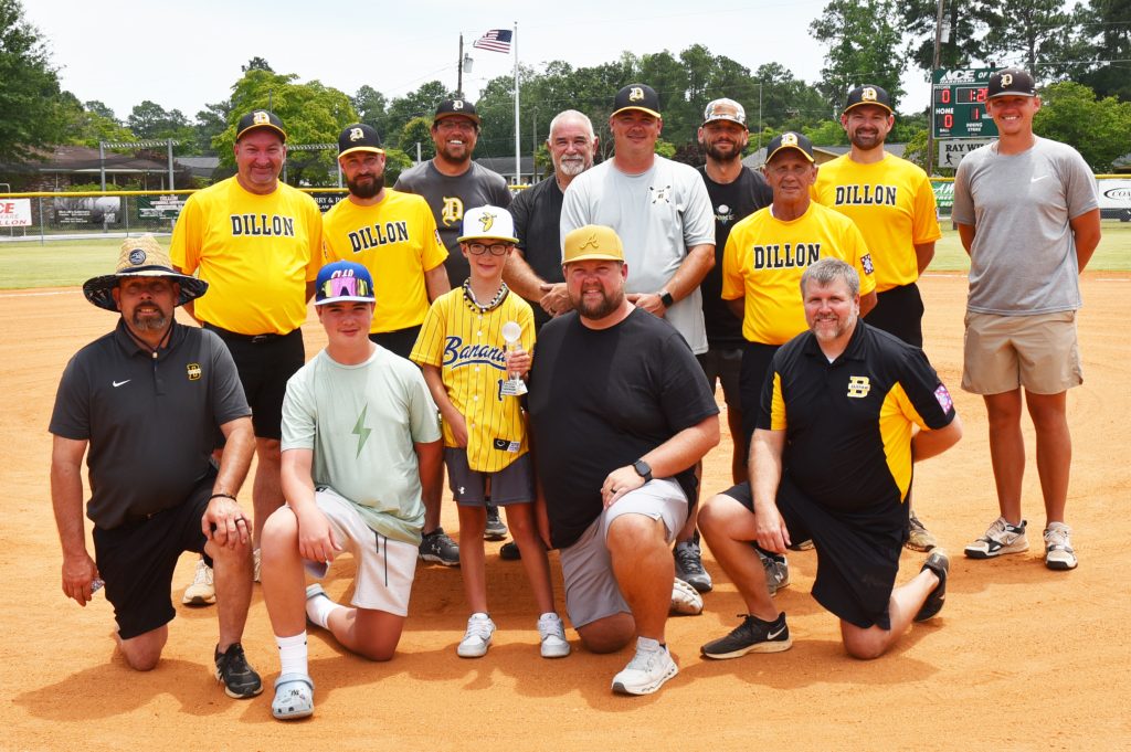 A group of adults and children pose together on a baseball field, some wearing yellow shirts labeled "DOLLOS." Trees and a flag are visible in the background.