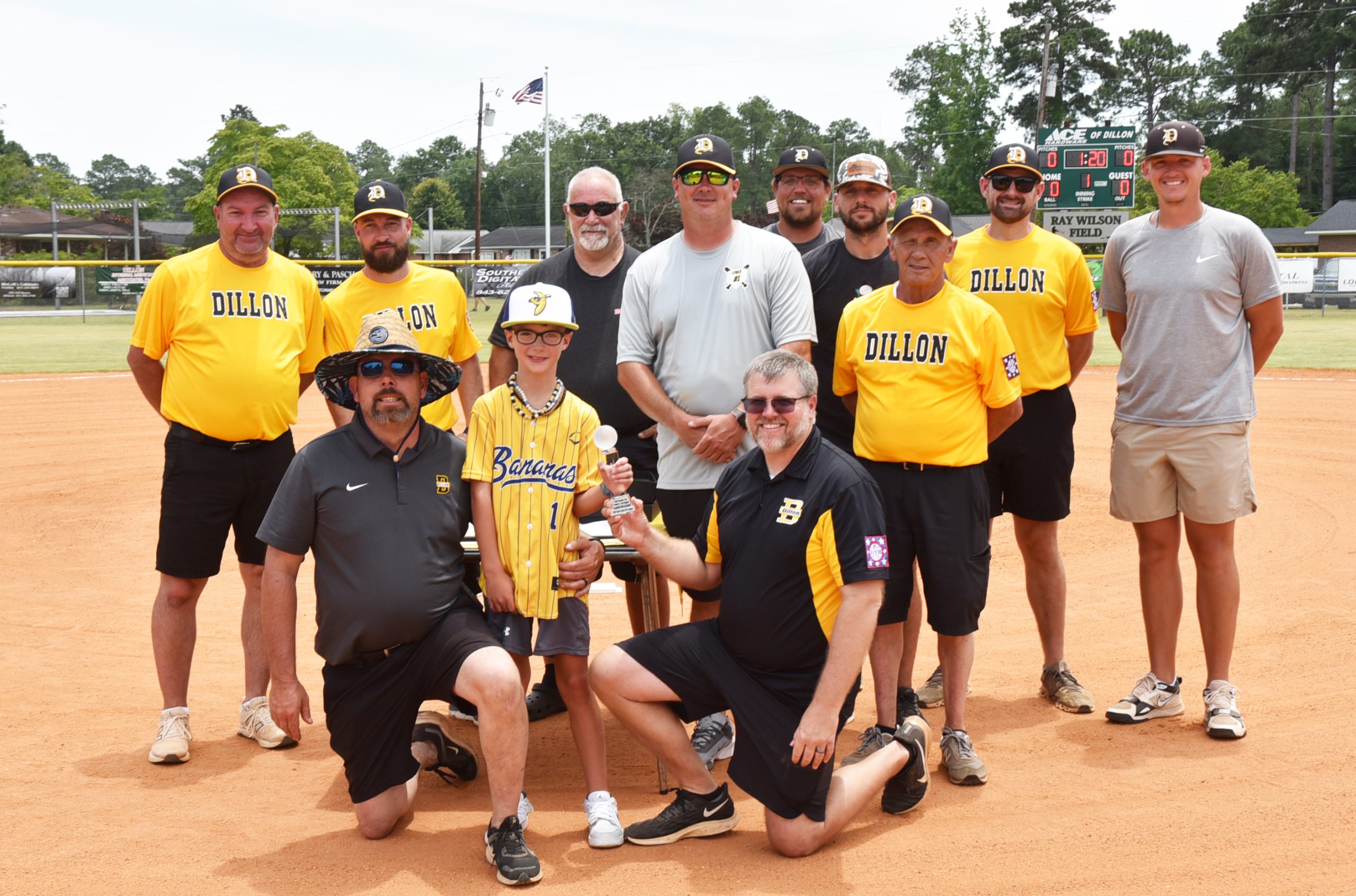 A youth baseball team and coaches pose on a field, some kneeling and some standing, all wearing matching yellow and black uniforms, smiling for the photo.
