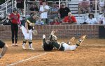Two softball players in green uniforms attempt to tag out a sliding runner at home plate as spectators watch from the stands.