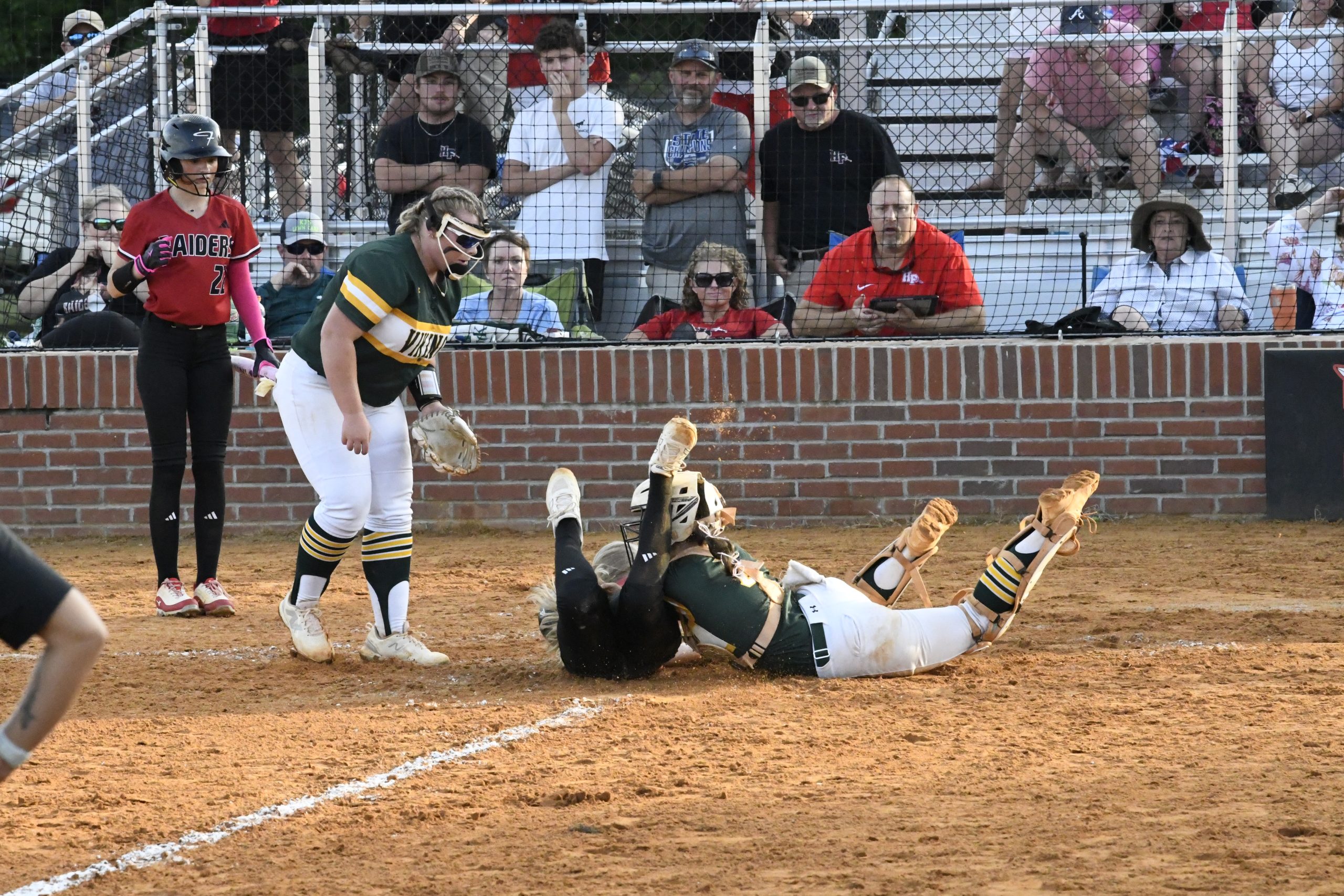 Two softball players in green uniforms attempt to tag out a sliding runner at home plate as spectators watch from the stands.