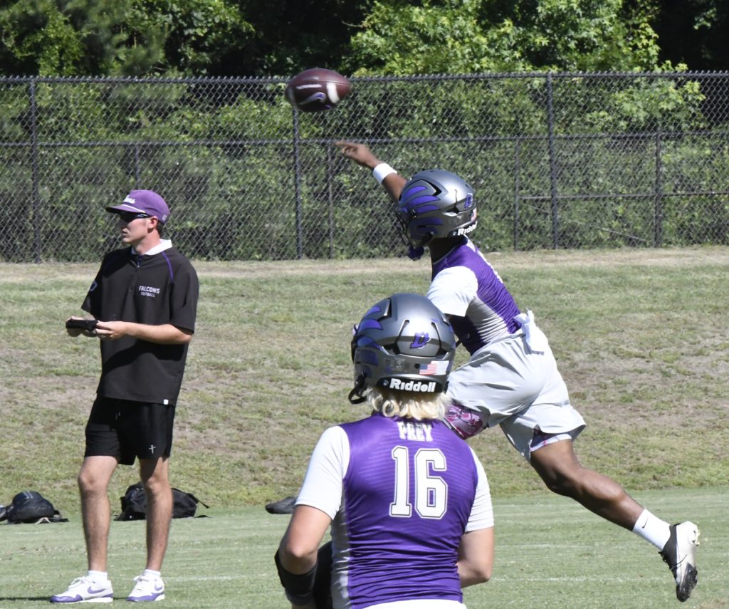 Football player in purple jersey throws a pass during practice, while a coach and another player watch on a grassy field.